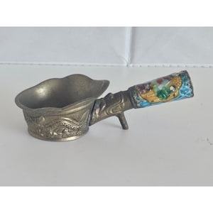 May include: A metallic ladle with a scalloped bowl featuring an embossed dragon design on the exterior. The handle is decorated with colorful enamel inlays depicting a figure and landscape, and includes a small metal support stand underneath.