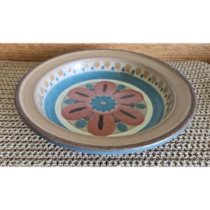 May include: A round ceramic bowl with a floral design. The bowl features a brown rim, a blue outer ring, and a central design with a large flower in shades of brown, teal, and blue. The bowl is on a textured surface.