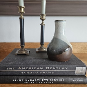 May include: A decorative arrangement featuring two brass and black candlesticks with lit candles, a small ceramic vase with brown, gray, and white tones, and two books titled "The American Century" and "Linda McCartney's Sixties."