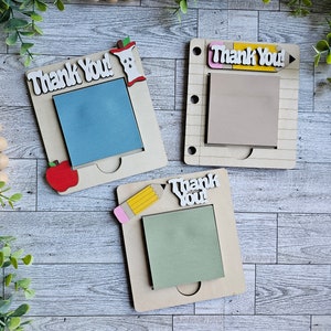 May include: Three wooden notepads with the words "Thank You!" on each. The notepads have different designs, including an apple, a pencil, and a pencil with a pink eraser. The notepads are on a light wood background.