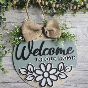 May include: Round wooden welcome sign with a burlap bow and rope hanger. The sign features the words "Welcome to our Home" in black lettering, with a daisy and leaf design at the bottom. Greenery and wooden beads surround the sign.