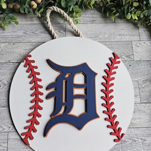 May include: A white wooden baseball-shaped door hanger with a blue and orange "D" in the center. The baseball has red stitching around the edges.