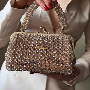 Può includere: Una borsa rettangolare dorata con perline, con chiusura a scatto e manico con perline. La borsa è ricoperta di piccole perline scintillanti. La parola "PARAGON" è visualizzata su una piccola placca dorata.