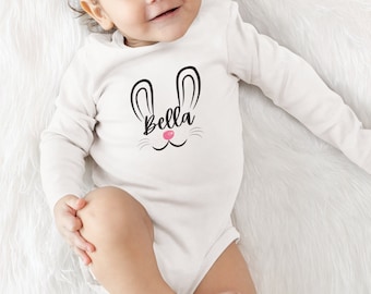 Könnte beinhalten: Ein weißer Langarmbody mit einem schwarzen Hasengesicht und dem Namen "Bella" auf der Vorderseite. Der Body wird von einem Baby getragen, das auf einer weißen, flauschigen Oberfläche liegt.
