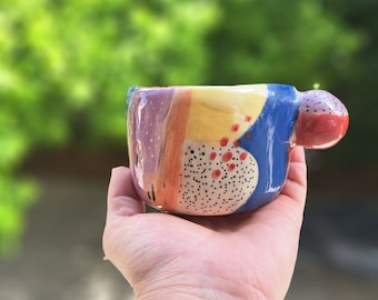 May include: A small, colorful ceramic mug with a unique abstract design. The mug features a yellow, blue, purple, and red color scheme with black and white polka dots. The mug has a handle that is shaped like a rounded knob.