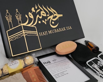 May include: A black gift box with gold lettering that says "Hajj Mubarak Isa" and a gold image of the Kaaba. The box is surrounded by various items, including a small bag with gold lettering, a wooden comb, a pair of scissors, a small bottle of oil, a black face mask, a wooden brush, a framed print with the word "Hajj" and a definition, and a small set of grooming tools.