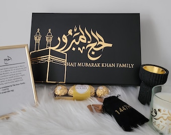May include: A black gift box with gold calligraphy that reads "Hajj Mubarak Khan Family". The box is surrounded by other gifts, including a framed print with information about the Hajj pilgrimage, a box of chocolates, a small pouch, a black incense burner, and a candle with gold calligraphy.
