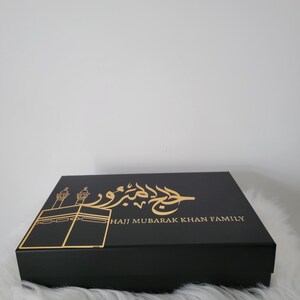 May include: Black gift box with gold lettering and a gold silhouette of the Kaaba. The text on the box reads "Hajj Mubarak Khan Family".