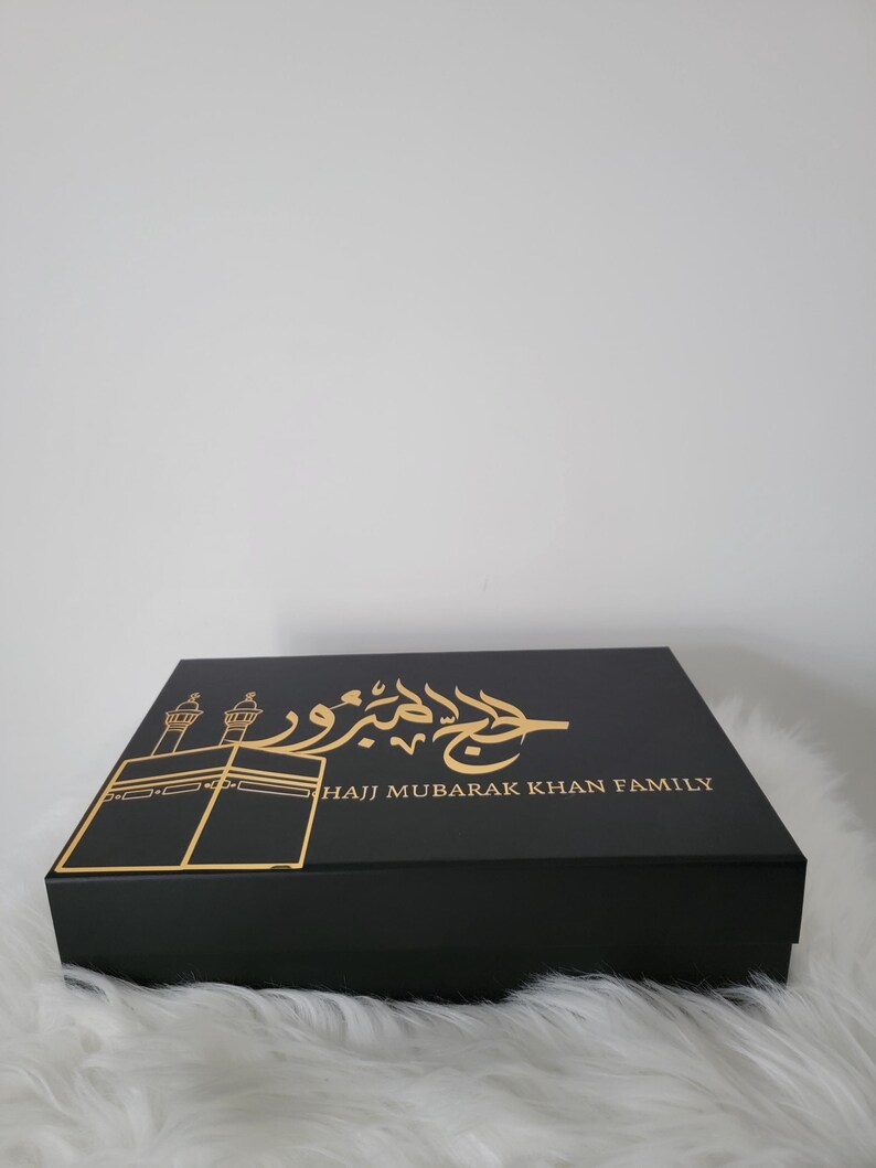 May include: Black gift box with gold lettering and a gold silhouette of the Kaaba. The text on the box reads "Hajj Mubarak Khan Family".