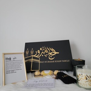 May include: A black gift box with gold lettering that says "Hajj Mubarak Khan Family". The box is filled with a variety of gifts, including chocolates, a candle, and a small black bag with gold lettering that says "1441".