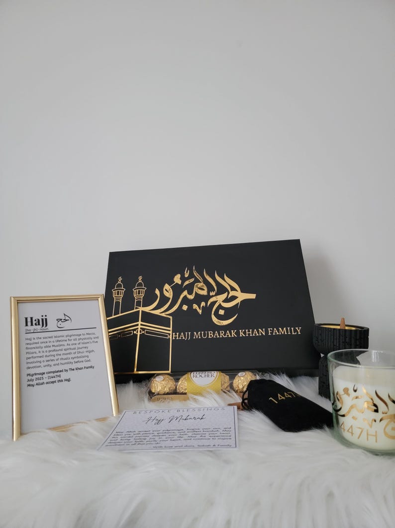 May include: A black gift box with gold lettering that says "Hajj Mubarak Khan Family". The box is filled with a variety of gifts, including chocolates, a candle, and a small black bag with gold lettering that says "1441".