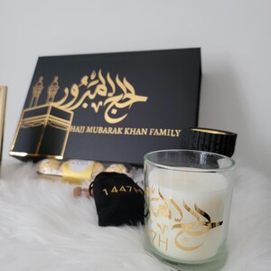 May include: A black gift box with gold lettering that reads "Hajj Mubarak Khan Family". The box is open and contains a small bag with gold lettering that reads "1447H", a box of chocolates, and a white candle with gold lettering in Arabic.