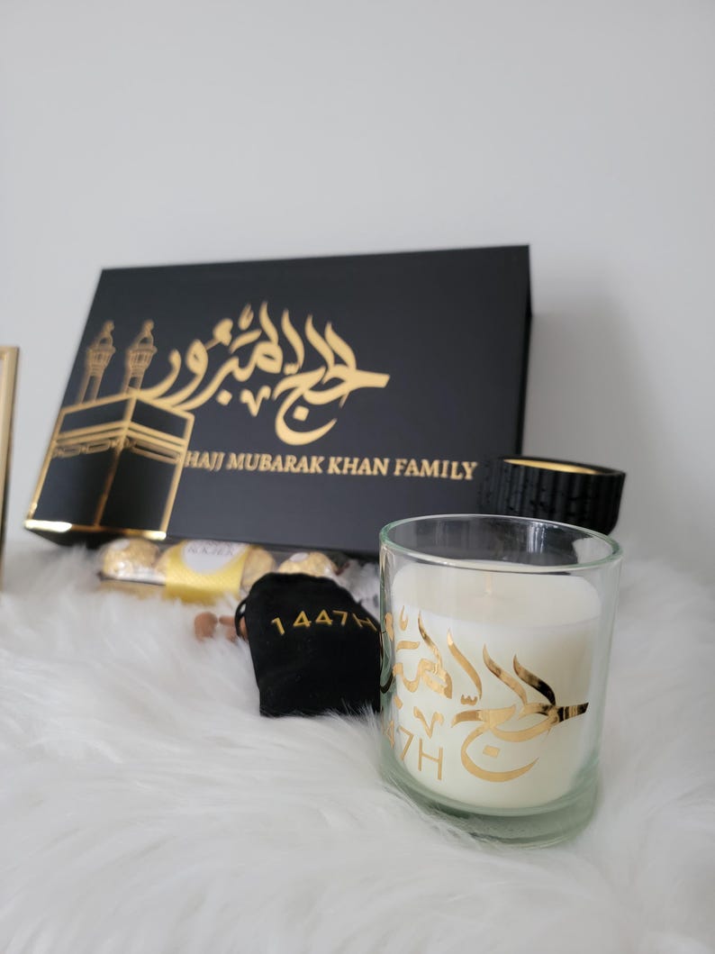 May include: A black gift box with gold lettering that reads "Hajj Mubarak Khan Family". The box is open and contains a small bag with gold lettering that reads "1447H", a box of chocolates, and a white candle with gold lettering in Arabic.