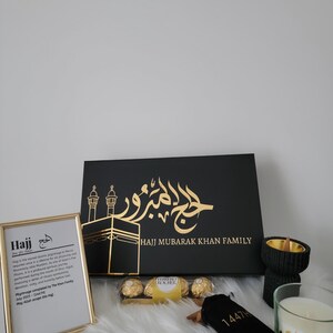 May include: A black gift box with gold lettering that says "Hajj Mubarak Khan Family". The box is decorated with a gold image of the Kaaba and a mosque. The box is surrounded by other gifts, including a framed print with the definition of Hajj, a bag of chocolates, a small black pouch with gold lettering, a black incense burner, and a white candle with gold lettering.