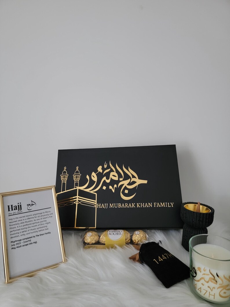 May include: A black gift box with gold lettering that says "Hajj Mubarak Khan Family". The box is decorated with a gold image of the Kaaba and a mosque. The box is surrounded by other gifts, including a framed print with the definition of Hajj, a bag of chocolates, a small black pouch with gold lettering, a black incense burner, and a white candle with gold lettering.