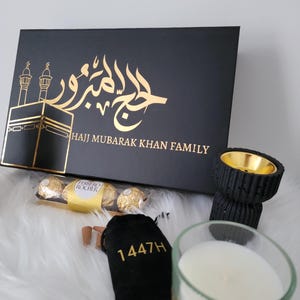 May include: A black gift box with gold lettering that reads "Hajj Mubarak Khan Family" and a gold Arabic calligraphy design. The box is surrounded by other gifts, including a black incense holder, a small bag of incense cones, a box of chocolates, and a candle with Arabic calligraphy.