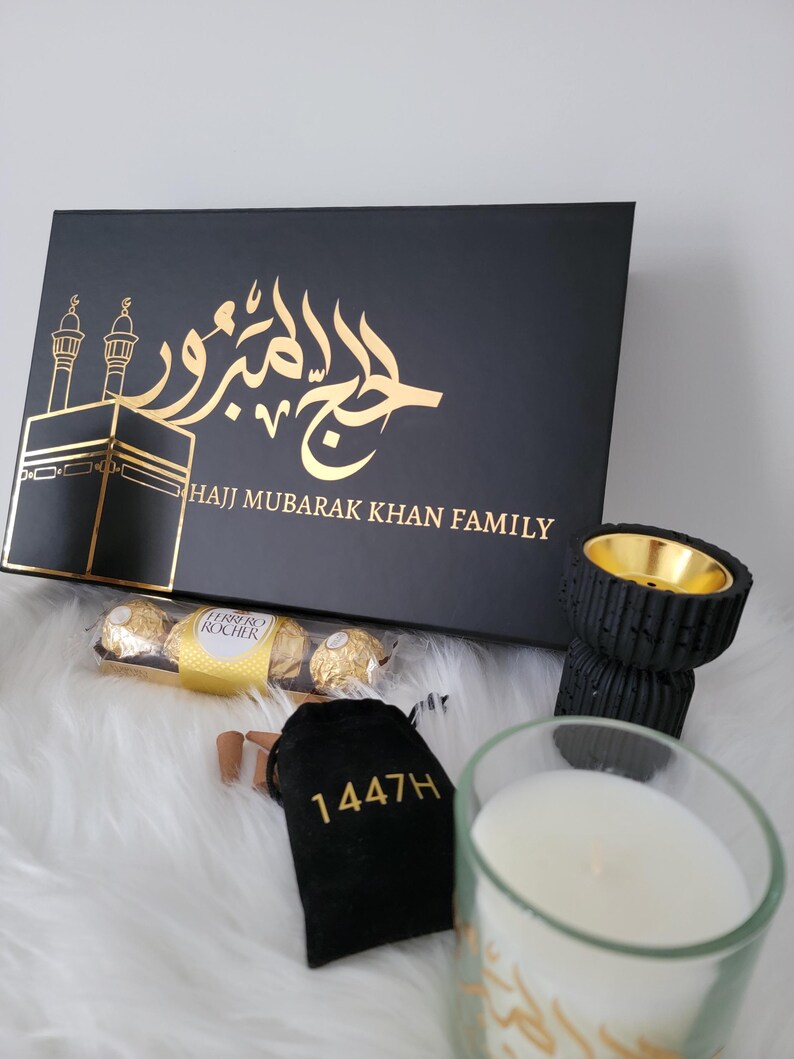 May include: A black gift box with gold lettering that reads "Hajj Mubarak Khan Family" and a gold Arabic calligraphy design. The box is surrounded by other gifts, including a black incense holder, a small bag of incense cones, a box of chocolates, and a candle with Arabic calligraphy.