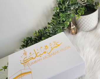 May include: White gift box with gold lettering that says "Umrah Mubarak Huda" and a gold illustration of the Kaaba.