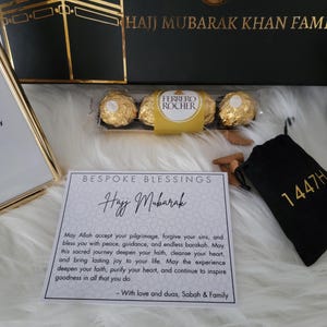 May include: A white card with gold lettering that reads "Bespoke Blessings Hajj Mubarak". The card has a message wishing someone a happy Hajj pilgrimage. The card is surrounded by gold chocolates, a small black bag with gold lettering that reads "1447H", and a small bag of incense sticks.