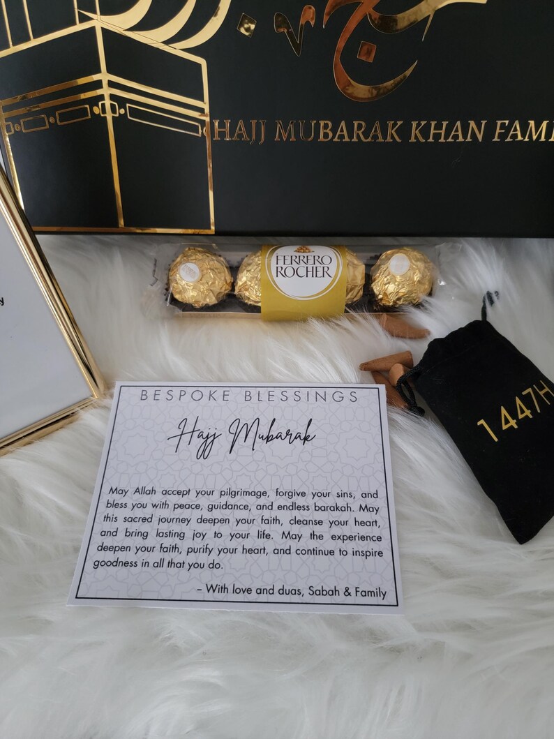 May include: A white card with gold lettering that reads "Bespoke Blessings Hajj Mubarak". The card has a message wishing someone a happy Hajj pilgrimage. The card is surrounded by gold chocolates, a small black bag with gold lettering that reads "1447H", and a small bag of incense sticks.