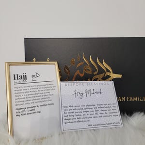 May include: A gold-framed picture with the word "Hajj" in black text and a definition of the word. A white card with gold lettering says "Bespoke Blessings Hajj Mubarak" and includes a message about the pilgrimage. The card is on a black surface.