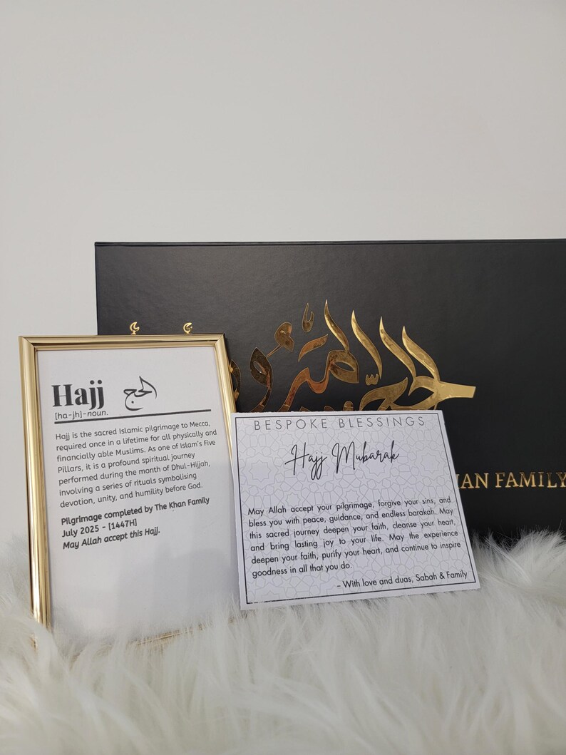 May include: A gold-framed picture with the word "Hajj" in black text and a definition of the word. A white card with gold lettering says "Bespoke Blessings Hajj Mubarak" and includes a message about the pilgrimage. The card is on a black surface.