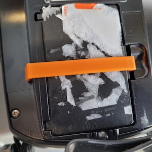 May include: Close-up of a black electronic device with an orange plastic latch. The device has a white sticker with the word "HERE" on it.