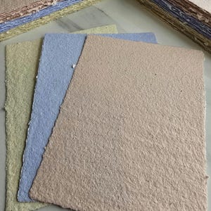 Può includere: Tre fogli di carta fatta a mano in diversi colori: verde chiaro, blu chiaro e marrone chiaro. La carta ha una superficie ruvida e testurizzata.