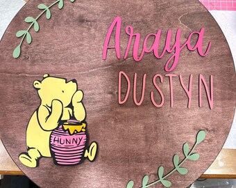 May include: A round wooden sign with the names "Araya Dustyn" written in pink script. A Winnie the Pooh character is holding a pink honey pot with the word "Hunny" on it. The sign is decorated with green leafy vines.