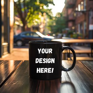 May include: A black ceramic coffee mug with a white inscription that reads "YOUR DESIGN HERE!" The mug is sitting on a wooden table.