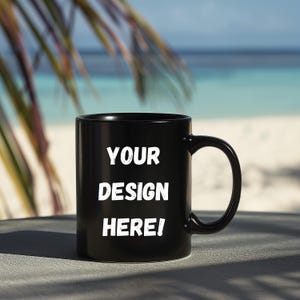 Puede incluir: Una taza de cerámica negra con el texto "YOUR DESIGN HERE!" en blanco. La taza tiene un asa curva y está colocada sobre un fondo de playa borroso con palmeras.