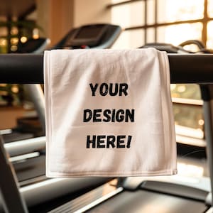 May include: White fitness towel with the text "YOUR DESIGN HERE!" in black. The towel is hanging on a black bar, with a treadmill and gym equipment in the background. The towel is made of a soft, absorbent material.