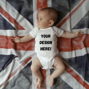 Op de afbeelding: Een witte baby romper met de tekst "YOUR DESIGN HERE!" op een rood, wit en blauwe Union Jack vlag.