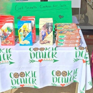 May include: A table displaying various boxes of Girl Scout cookies for sale. The table is covered with a white tablecloth printed with the words "Cookie Dealer" and cookie graphics. A green sign reads "Girl Scouts Cookies For Sale."