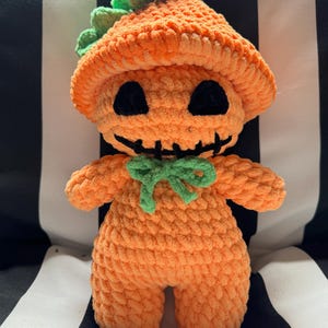 May include: A crocheted pumpkin-shaped plush toy with a green stem and a friendly face. The toy has black eyes, a stitched smile, and a green bow.