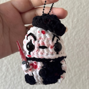 May include: Crocheted character with black beret, black eyes, and a red chainsaw. The character is white with black accents and red blood-like splatters. A black beaded chain is attached to the top.
