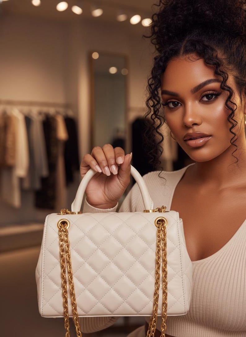 May include: Cream-colored quilted handbag with gold-tone chain straps and hardware. The bag has a top handle and a rectangular shape. The background shows a clothing store with racks of clothing.