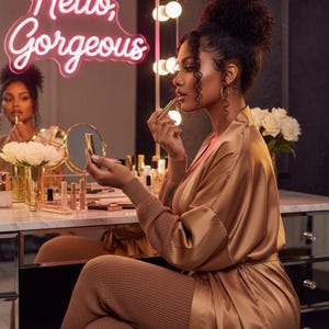 May include: A woman applying lipstick in front of a vanity mirror. The mirror has a neon sign that says "Hello, Gorgeous." She is wearing a gold satin robe and matching knit pants. The vanity is filled with makeup products and a vase of white roses.