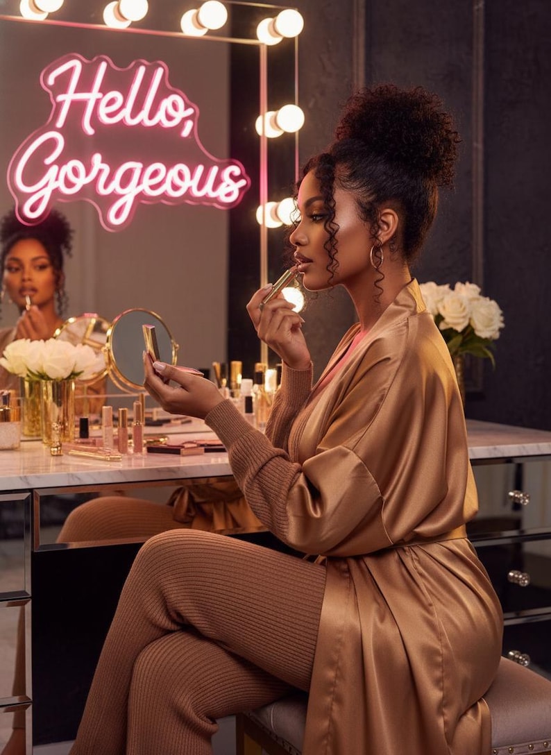 May include: A woman applying lipstick in front of a vanity mirror. The mirror has a neon sign that says "Hello, Gorgeous." She is wearing a gold satin robe and matching knit pants. The vanity is filled with makeup products and a vase of white roses.