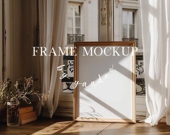 May include: A gold-colored frame mockup with a white background, sitting on a wooden floor in front of a window with white curtains. The frame is in a room with a vase of dried flowers and a stack of boxes.