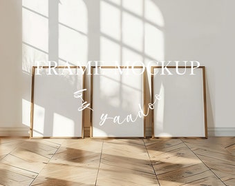 May include: Three gold picture frames with white mats are on a wooden floor. The frames are empty and ready for your artwork or photos. The floor is made of light wood planks with a chevron pattern. The frames are in a room with white walls and a window with sunlight streaming in.