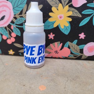 Puede incluir: Un pequeño frasco transparente de gotas para los ojos con una tapa blanca y el texto "BYE BYE PINK EYE" en azul. El frasco se encuentra sobre un fondo de un estampado floral negro con flores rosas y amarillas.