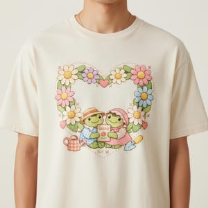 Può includere: T-shirt color crema con un design a forma di ghirlanda floreale a cuore. All'interno del cuore, due personaggi dei cartoni animati tengono in mano un pacchetto di semi. Sono raffigurati anche un annaffiatoio e una cazzuola. La maglietta ha girocollo e maniche corte.