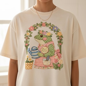 Può includere: T-shirt color crema con un design a forma di rana stravagante. La rana indossa un grembiule con la scritta "Plant Parent" e tiene un annaffiatoio. Il design include fiori e un arco floreale, creando un'estetica affascinante a tema giardino.