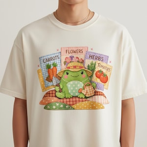 May include: Cream-colored t-shirt featuring a whimsical garden scene. A smiling frog in a straw hat is surrounded by illustrated signs for carrots, flowers, herbs, and tomatoes. The design includes a watering can and a shovel, with a patchwork quilt base.
