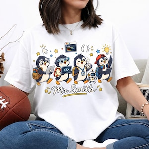 May include: A white t-shirt featuring a graphic of four cartoon penguins engaged in tech-related activities, including using laptops and a small robot. The text "Mr. Smith" is printed in a cursive font below the penguins, surrounded by stars.