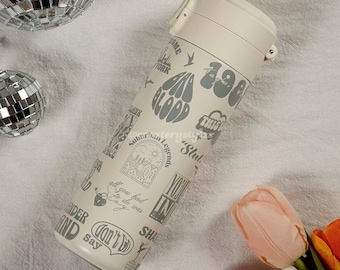 May include: A beige stainless steel water bottle with a gray print of various phrases and designs, including "Bad Blood", "Suburban Legends", "Slut", "You had to do was crazy", "Can't Stop", and "This Was a House".