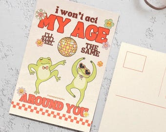 Puede incluir: Una postal con el texto "I won't act my age, I'll still feel the same around you" e ilustraciones de ranas bailando, una bola de discoteca y flores. La postal tiene un diseño retro con un borde a cuadros.