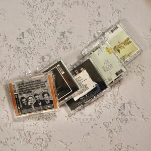 May include: Four clear plastic CD cases with different album covers. The covers feature a band with four members, a band with one member, a band with three members, and a band with two members. The cases are stacked on top of each other.