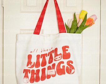 May include: White canvas tote bag with red straps and the text "all these little things" in red stylized lettering. The bag has a red heart, a red cassette tape, a red cup of coffee, and a red mouth graphic.
